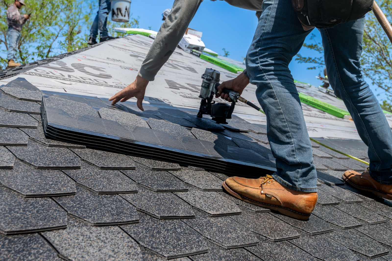 A roofer using a nail gun to lay asphalt shingles over roof underlayment